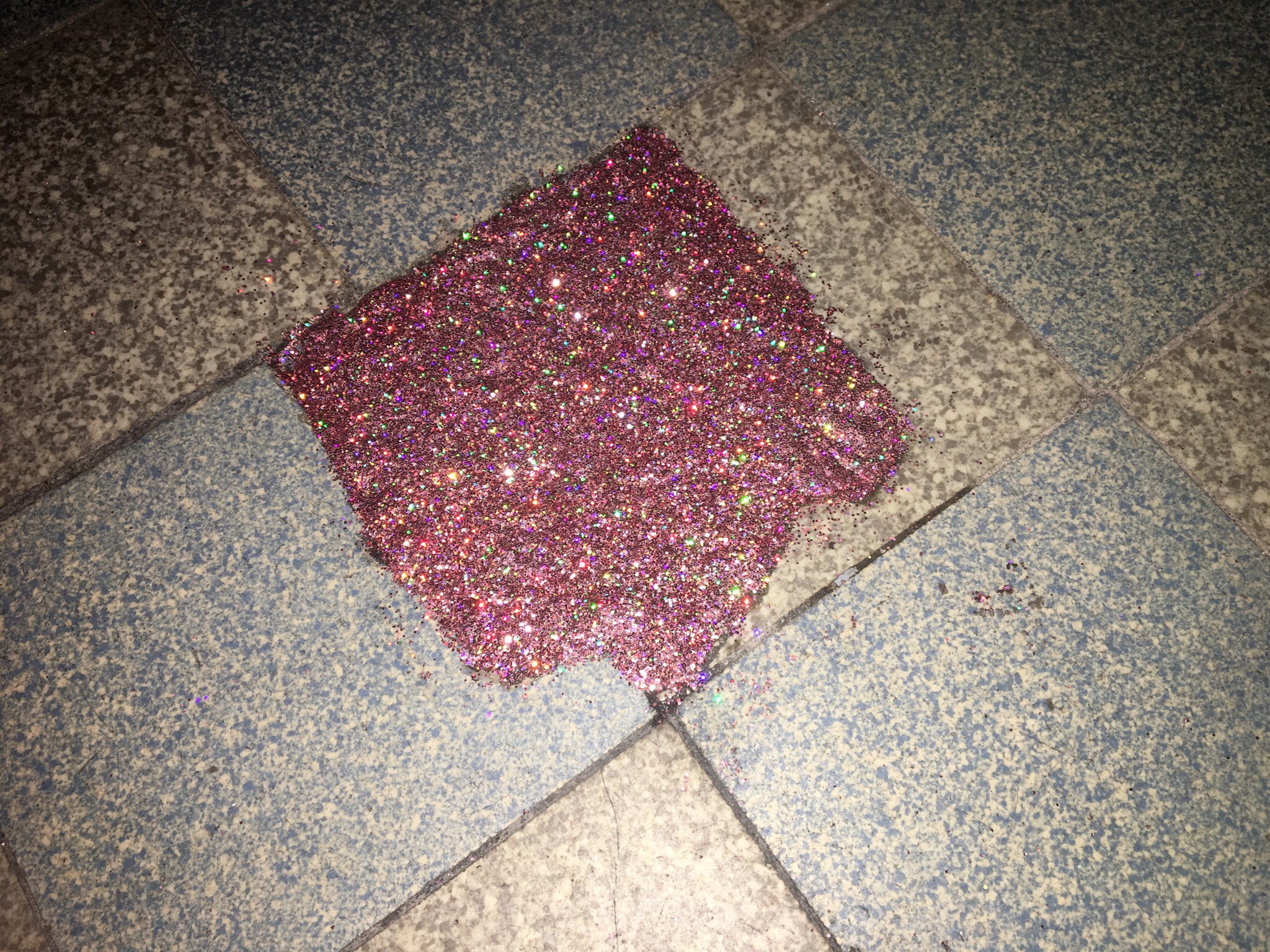 Un sol de carrelage est abimé, il semble manquer un carreau. Il a été réparé avec un applat de paillettes roses. Photo avec flash qui fait plus ressortir les pailletes qui sont holographiques.